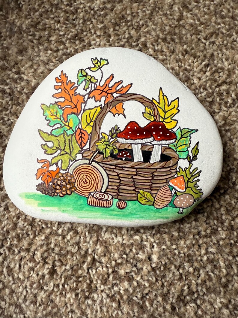 Puede incluir: Una piedra blanca pintada a mano presenta una cesta marr&oacute;n llena de setas con gorro rojo y follaje verde. Hojas de oto&ntilde;o en naranja, amarillo y verde rodean la cesta. Detalles pintados adicionales: caracol, uvas y otras setas.