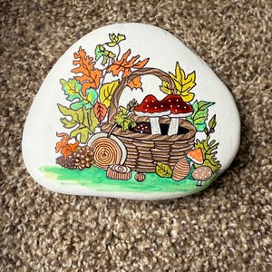 Puede incluir: Una piedra blanca lisa pintada a mano presenta un dise&ntilde;o con tem&aacute;tica oto&ntilde;al. Una cesta tejida est&aacute; llena de setas y hojas en tonos rojos, naranjas, amarillos y verdes. Detalles adicionales incluyen rodajas de madera y una base verde.