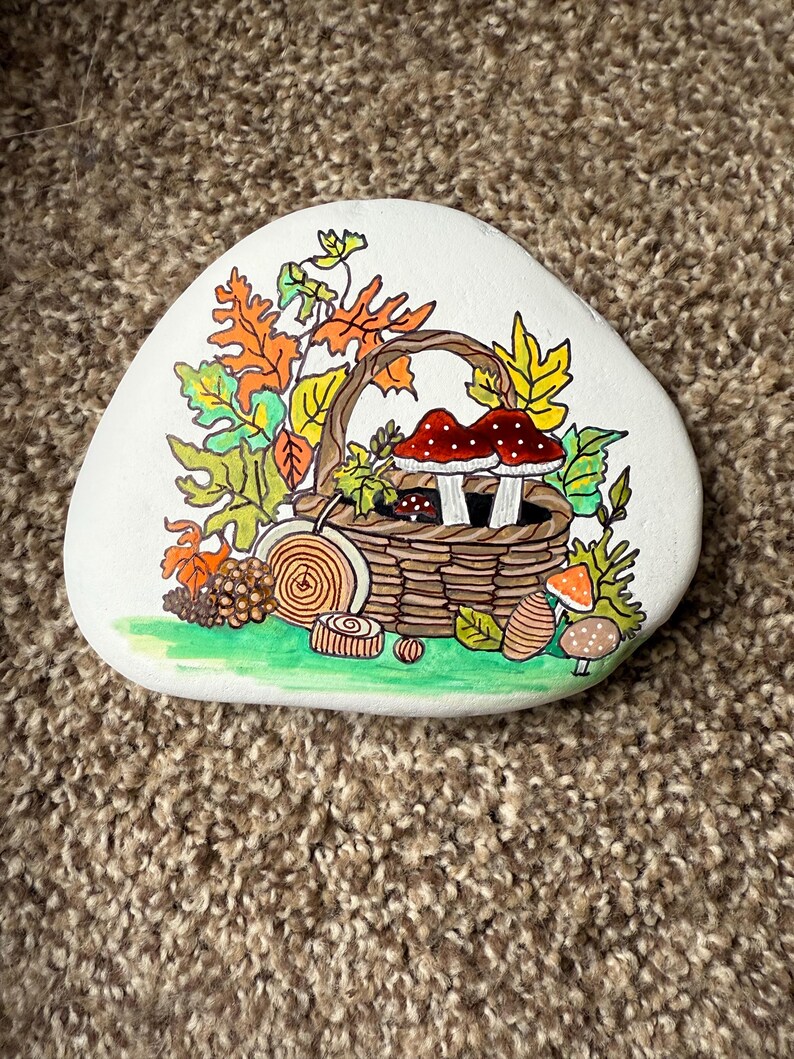 Puede incluir: Una piedra blanca lisa pintada a mano presenta un dise&ntilde;o con tem&aacute;tica oto&ntilde;al. Una cesta tejida est&aacute; llena de setas y hojas en tonos rojos, naranjas, amarillos y verdes. Detalles adicionales incluyen rodajas de madera y una base verde.