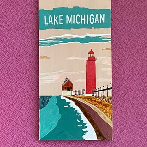 K&ouml;nnte beinhalten: Ein Holzschild mit dem Text "LAKE MICHIGAN" in wei&szlig; auf einem t&uuml;rkisfarbenen Hintergrund. Das Schild zeigt einen roten Leuchtturm mit einem wei&szlig;en Dach und einem wei&szlig;en Zaun auf einem braunen Steg. Der Steg erstreckt sich in ein t&uuml;rkisfarbenes Gew&auml;sser mit wei&szlig;en Wellen.