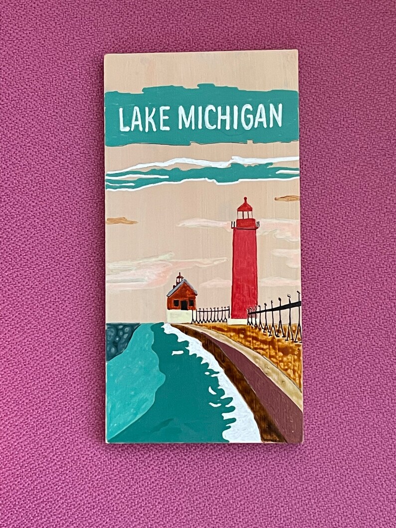 K&ouml;nnte beinhalten: Ein Holzschild mit dem Text "LAKE MICHIGAN" in wei&szlig; auf einem t&uuml;rkisfarbenen Hintergrund. Das Schild zeigt einen roten Leuchtturm mit einem wei&szlig;en Dach und einem wei&szlig;en Zaun auf einem braunen Steg. Der Steg erstreckt sich in ein t&uuml;rkisfarbenes Gew&auml;sser mit wei&szlig;en Wellen.