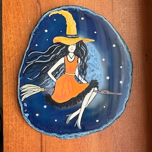 Puede incluir: Una bruja pintada a mano en una escoba, sobre un fondo azul oscuro, estilo ágata. La bruja lleva un vestido naranja y un sombrero de bruja amarillo. La obra de arte está sobre una piedra decorativa de forma ovalada.