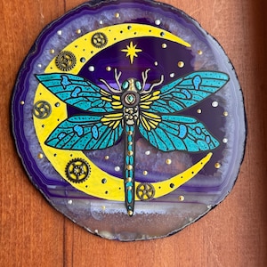 Puede incluir: Una rodaja de ágata decorativa y redonda con un diseño de libélula pintada. La libélula tiene alas turquesas y un cuerpo amarillo, sobre una luna creciente amarilla y un fondo morado oscuro. Engranajes y estrellas doradas añaden detalles.