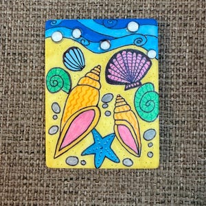 May include: A rectangular painting featuring a beach scene with a yellow background, blue waves, and various seashells. The seashells are in shades of orange, pink, green, and blue, with a blue starfish. Small gray stones are scattered around the seashells.