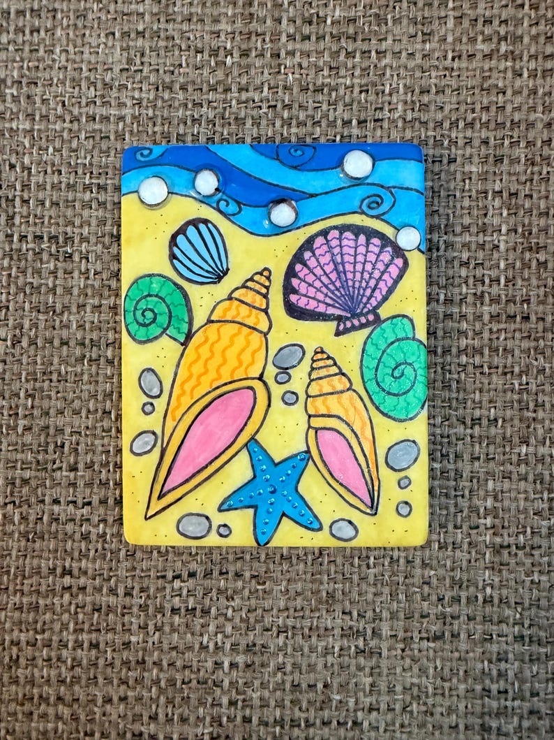 May include: A rectangular painting featuring a beach scene with a yellow background, blue waves, and various seashells. The seashells are in shades of orange, pink, green, and blue, with a blue starfish. Small gray stones are scattered around the seashells.