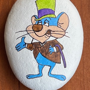 Puede incluir: Pintura de roca pintada a mano que representa a un rat&oacute;n de dibujos animados con un sombrero de copa verde y morado, una chaqueta marr&oacute;n y pantalones azules. El rat&oacute;n sostiene un bast&oacute;n en su mano derecha y tiene una expresi&oacute;n de sorpresa en su rostro.