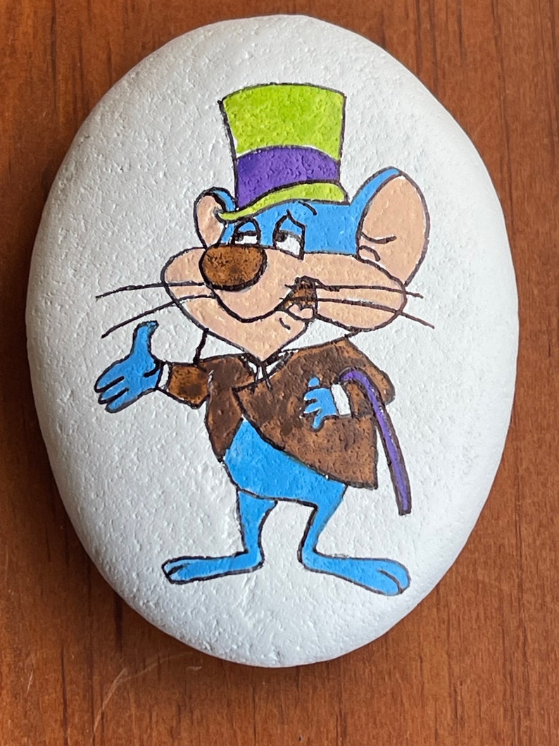 Puede incluir: Pintura de roca pintada a mano que representa a un rat&oacute;n de dibujos animados con un sombrero de copa verde y morado, una chaqueta marr&oacute;n y pantalones azules. El rat&oacute;n sostiene un bast&oacute;n en su mano derecha y tiene una expresi&oacute;n de sorpresa en su rostro.