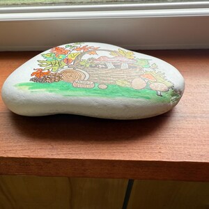 Puede incluir: Una piedra blanca lisa pintada a mano con una escena oto&ntilde;al caprichosa. La obra de arte representa una casa de setas, hojas coloridas y varias setas sobre una zona de hierba verde. La piedra est&aacute; sobre una superficie de madera.