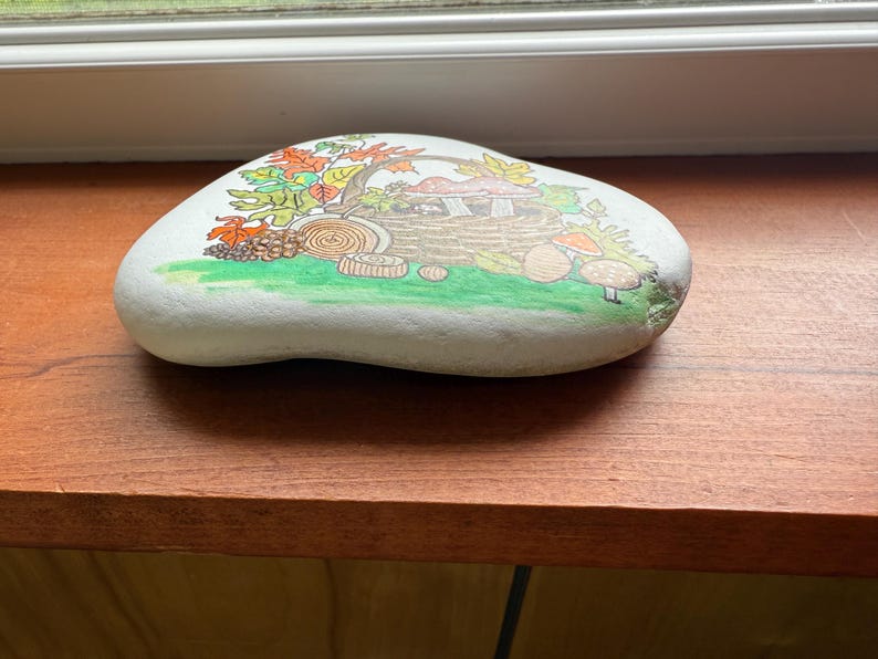 Puede incluir: Una piedra blanca lisa pintada a mano con una escena oto&ntilde;al caprichosa. La obra de arte representa una casa de setas, hojas coloridas y varias setas sobre una zona de hierba verde. La piedra est&aacute; sobre una superficie de madera.