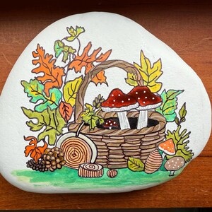 Puede incluir: Piedra pintada a mano con tem&aacute;tica oto&ntilde;al. Una cesta tejida contiene dos setas de sombrero rojo con lunares blancos. La cesta est&aacute; rodeada de hojas coloridas, una pi&ntilde;a y otros elementos decorativos. La piedra est&aacute; pintada sobre un fondo blanco.