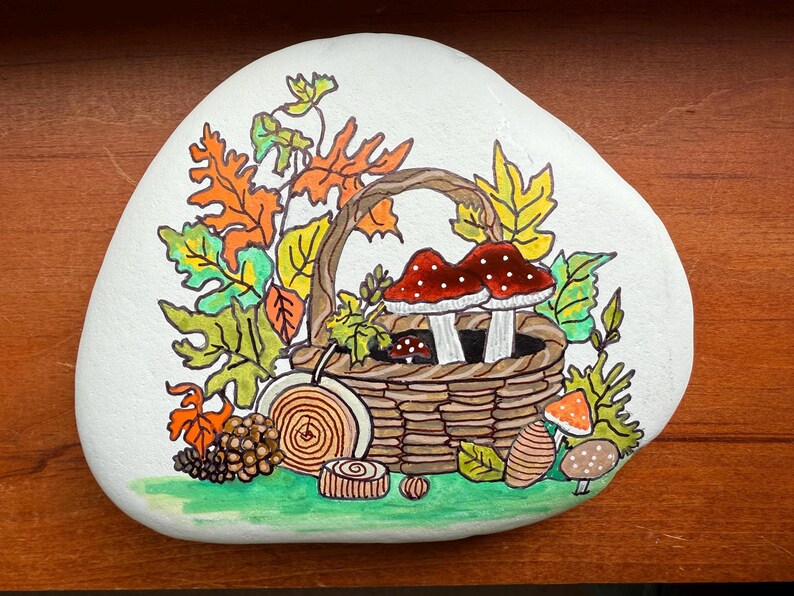 Puede incluir: Piedra pintada a mano con tem&aacute;tica oto&ntilde;al. Una cesta tejida contiene dos setas de sombrero rojo con lunares blancos. La cesta est&aacute; rodeada de hojas coloridas, una pi&ntilde;a y otros elementos decorativos. La piedra est&aacute; pintada sobre un fondo blanco.