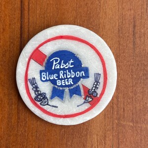 Puede incluir: Un posavasos redondo blanco con un c&iacute;rculo rojo y una l&iacute;nea roja diagonal que lo atraviesa. Dentro del c&iacute;rculo hay un logotipo azul de Pabst Blue Ribbon Beer.