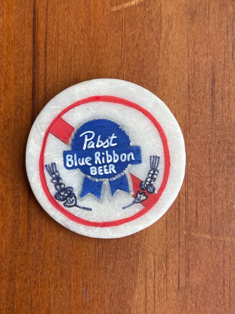 Puede incluir: Un posavasos redondo blanco con un c&iacute;rculo rojo y una l&iacute;nea roja diagonal que lo atraviesa. Dentro del c&iacute;rculo hay un logotipo azul de Pabst Blue Ribbon Beer.