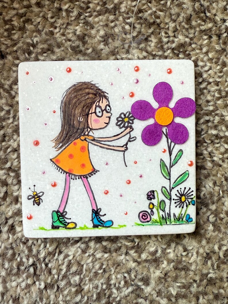 Puede incluir: Una ilustraci&oacute;n caprichosa en un cuadrado blanco representa a una ni&ntilde;a con gafas con un vestido naranja, recogiendo una flor. Tambi&eacute;n se presenta una gran flor de fieltro p&uacute;rpura y una abeja. Los puntos rojos y naranjas a&ntilde;aden un toque l&uacute;dico.