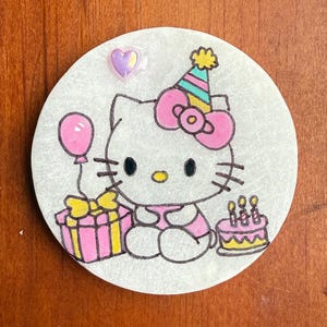 Op de afbeelding: Een ronde witte keramische ornament met een roze Hello Kitty-figuur die een feestmuts draagt, een roze cadeaudoos en een roze ballon vasthoudt, en een roze en gele verjaardagstaart met kaarsen.
