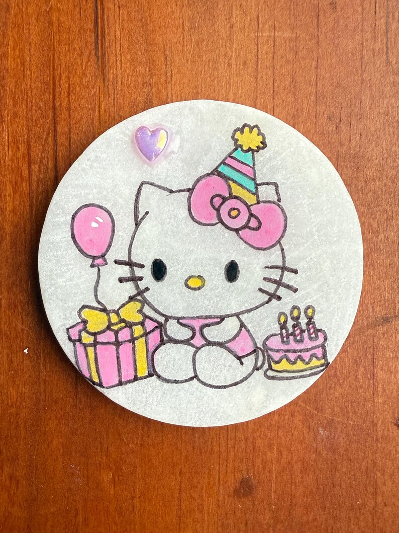Op de afbeelding: Een ronde witte keramische ornament met een roze Hello Kitty-figuur die een feestmuts draagt, een roze cadeaudoos en een roze ballon vasthoudt, en een roze en gele verjaardagstaart met kaarsen.