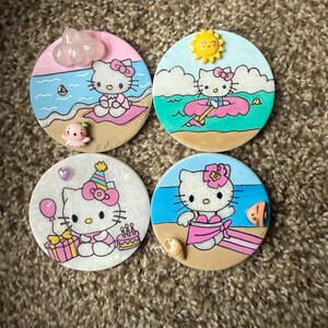 Op de afbeelding: Vier ronde keramische onderzetters met Hello Kitty in verschillende zomerse sc&egrave;nes. De onderzetters zijn geschilderd met Hello Kitty in een roze badpak, een roze zwemband, een verjaardagshoed en een roze bikini. Elke onderzetter heeft een andere achtergrondsc&egrave;ne.