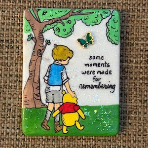 May include: A rectangular plaque featuring a hand-drawn illustration of a child and a yellow bear walking near a tree. The text reads, some moments were made for remembering. A small green butterfly charm is attached, and the surface has glitter.