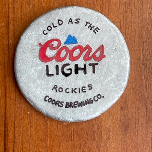 Puede incluir: Una tapa de botella de metal plateado con las palabras "Cold as the Coors Light Rockies Coors Brewing Co." impresas en ella.