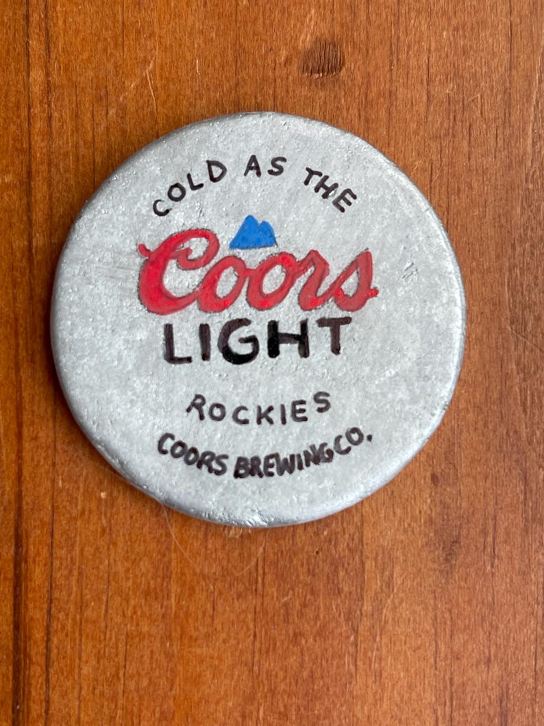 Puede incluir: Una tapa de botella de metal plateado con las palabras "Cold as the Coors Light Rockies Coors Brewing Co." impresas en ella.