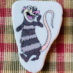 May include: Hand-painted rock art featuring a cartoon possum with a pink tail, black and white stripes, and a wide open mouth. The possum is painted on a white, oval-shaped rock.
