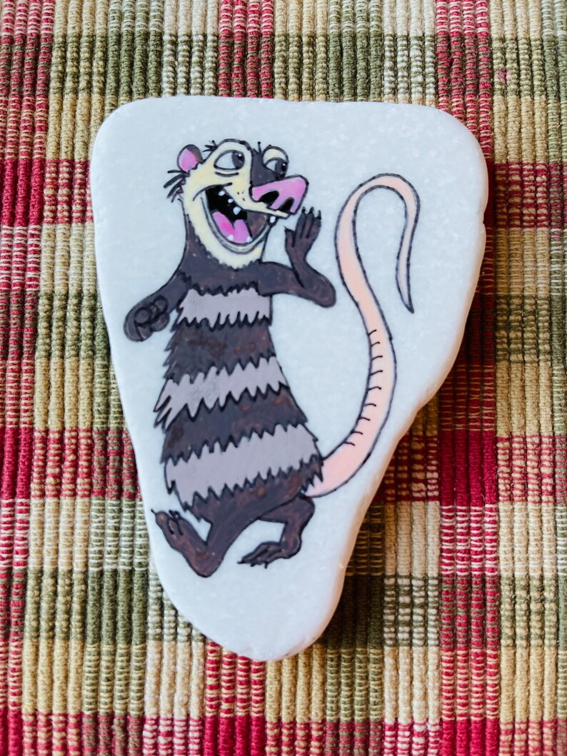May include: Hand-painted rock art featuring a cartoon possum with a pink tail, black and white stripes, and a wide open mouth. The possum is painted on a white, oval-shaped rock.