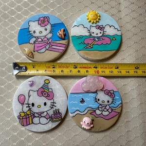 Op de afbeelding: Vier handgemaakte keramische onderzetters met Hello Kitty in verschillende zomerse sc&egrave;nes. De onderzetters zijn rond en hebben een witte achtergrond met kleurrijke details. De eerste onderzetter toont Hello Kitty in een roze bikini op een strand met een schelp en een surfboard. De tweede onderzetter toont Hello Kitty in een blauw shirt en roze shorts op een roze drijver in de oceaan. De derde onderzetter toont Hello Kitty in een roze jurk en een feestmuts die een ballon en een cadeau vasthoudt. De vierde onderzetter toont Hello Kitty in een roze badjas die op een handdoek op het strand zit met een roze octopus.