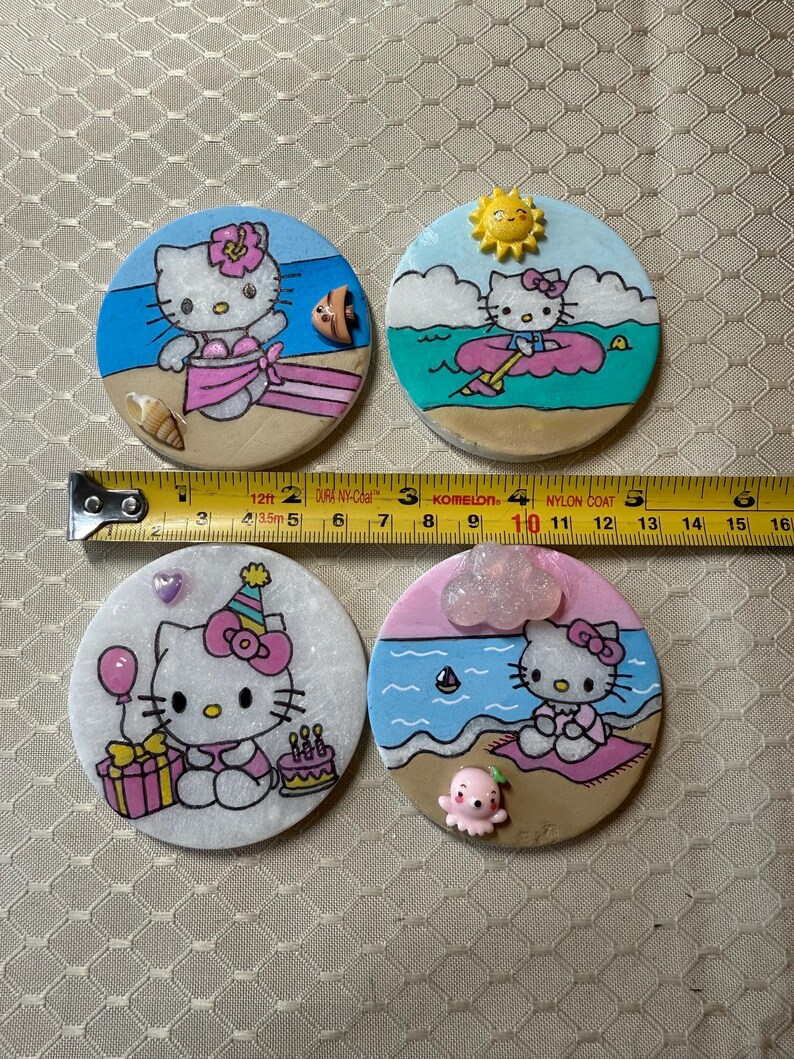 Op de afbeelding: Vier handgemaakte keramische onderzetters met Hello Kitty in verschillende zomerse sc&egrave;nes. De onderzetters zijn rond en hebben een witte achtergrond met kleurrijke details. De eerste onderzetter toont Hello Kitty in een roze bikini op een strand met een schelp en een surfboard. De tweede onderzetter toont Hello Kitty in een blauw shirt en roze shorts op een roze drijver in de oceaan. De derde onderzetter toont Hello Kitty in een roze jurk en een feestmuts die een ballon en een cadeau vasthoudt. De vierde onderzetter toont Hello Kitty in een roze badjas die op een handdoek op het strand zit met een roze octopus.