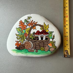 Puede incluir: Una piedra blanca pintada a mano con un dise&ntilde;o oto&ntilde;al. Una cesta marr&oacute;n llena de setas est&aacute; rodeada de hojas coloridas. La piedra mide aproximadamente 13 cm de largo, con una cinta m&eacute;trica amarilla en el fondo.