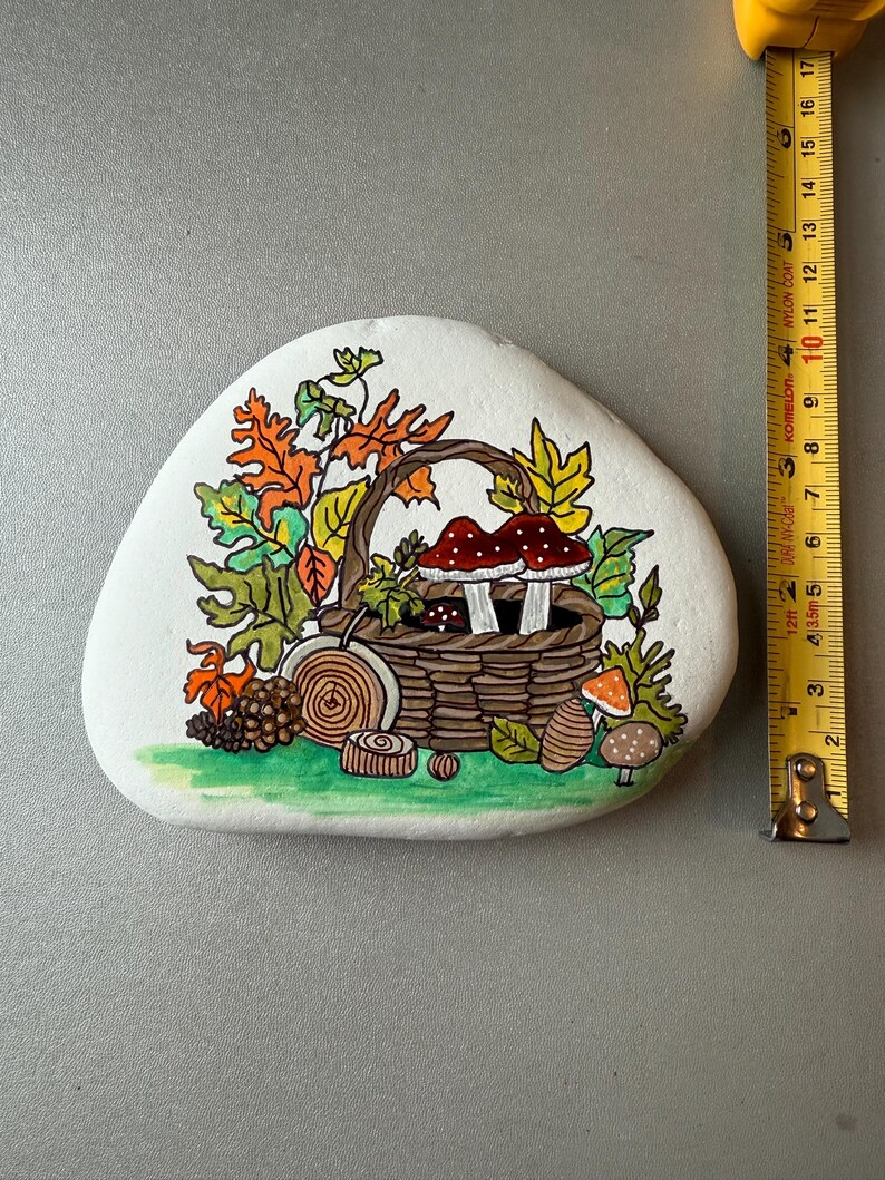 Puede incluir: Una piedra blanca pintada a mano con un dise&ntilde;o oto&ntilde;al. Una cesta marr&oacute;n llena de setas est&aacute; rodeada de hojas coloridas. La piedra mide aproximadamente 13 cm de largo, con una cinta m&eacute;trica amarilla en el fondo.