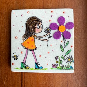 Puede incluir: Azulejo de cer&aacute;mica cuadrado con una ilustraci&oacute;n de dibujos animados de una ni&ntilde;a con un vestido naranja recogiendo una flor. A la derecha hay una gran flor morada con un centro naranja. Tambi&eacute;n se representa una abeja y otras flores.