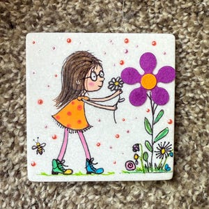 Puede incluir: Azulejo de cer&aacute;mica cuadrado con una ilustraci&oacute;n caprichosa de una ni&ntilde;a con un vestido naranja y gafas, recogiendo una margarita. Una gran flor morada con un centro naranja est&aacute; a su lado. Tambi&eacute;n se representa una abeja y otras flores.