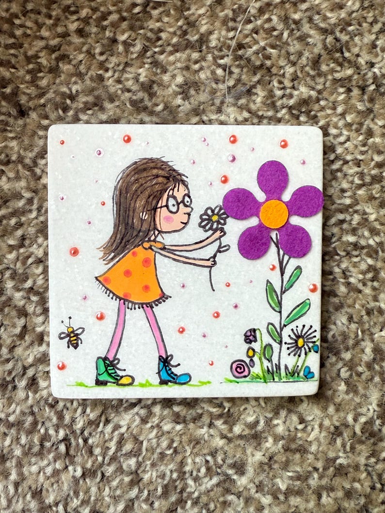 Puede incluir: Azulejo de cer&aacute;mica cuadrado con una ilustraci&oacute;n caprichosa de una ni&ntilde;a con un vestido naranja y gafas, recogiendo una margarita. Una gran flor morada con un centro naranja est&aacute; a su lado. Tambi&eacute;n se representa una abeja y otras flores.