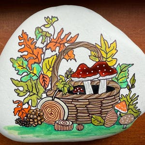 Puede incluir: Una piedra blanca pintada a mano con un dise&ntilde;o oto&ntilde;al. Una cesta tejida contiene dos setas de sombrero rojo con puntos blancos. El dise&ntilde;o incluye hojas coloridas, una pi&ntilde;a y otros elementos oto&ntilde;ales.