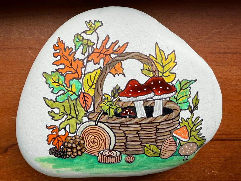 Puede incluir: Una piedra blanca pintada a mano con un dise&ntilde;o oto&ntilde;al. Una cesta tejida contiene dos setas de sombrero rojo con puntos blancos. El dise&ntilde;o incluye hojas coloridas, una pi&ntilde;a y otros elementos oto&ntilde;ales.