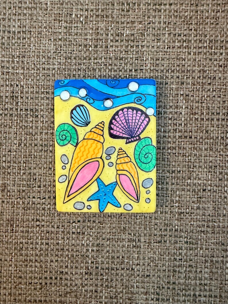 May include: A rectangular, hand-painted ceramic tile featuring a beach scene. The design includes yellow sand, blue waves, and various seashells in shades of orange, pink, and green. A blue starfish and small gray pebbles complete the coastal theme.