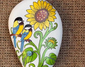 May include: A white, teardrop-shaped stone painted with a large yellow sunflower, two small birds with yellow and blue feathers perched on a green stem, a green leaf, a closed flower bud, and a small light blue ladybird.