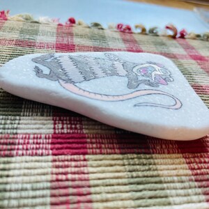 May include: Hand-painted rock with a gray and white striped opossum with pink accents. The opossum is holding a pink heart and has a pink nose.