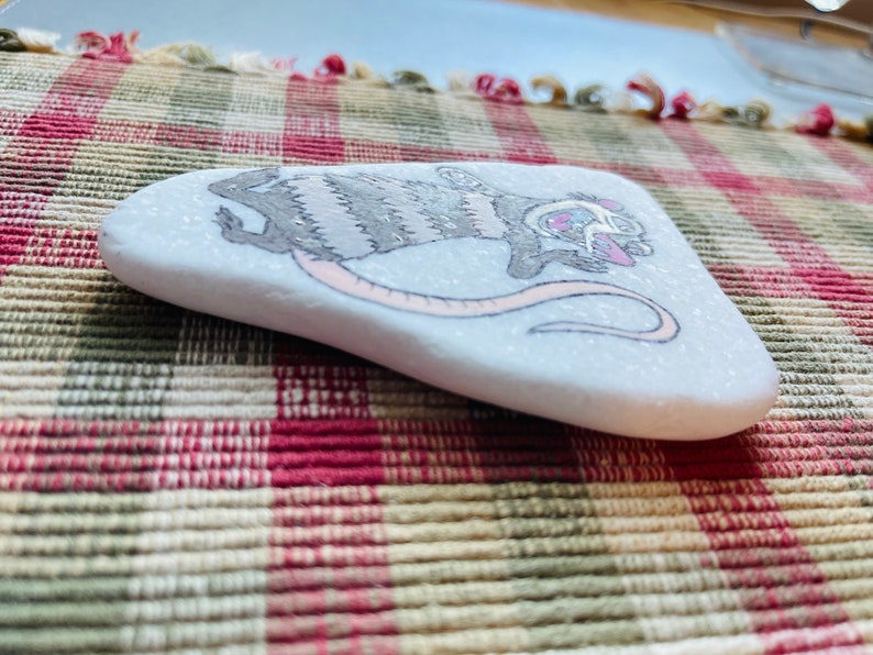 May include: Hand-painted rock with a gray and white striped opossum with pink accents. The opossum is holding a pink heart and has a pink nose.