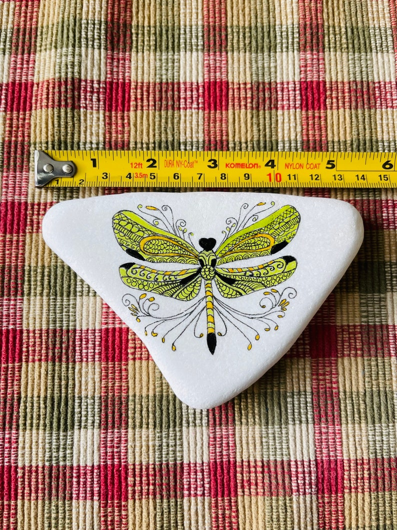 K&ouml;nnte beinhalten: Ein wei&szlig;er dreieckiger Stein, der mit einer gr&uuml;nen und gelben Libelle mit schwarzen Umrissen bemalt ist. Die Libelle hat komplizierte Details und ein schwarzes Herz in der Mitte ihres K&ouml;rpers.