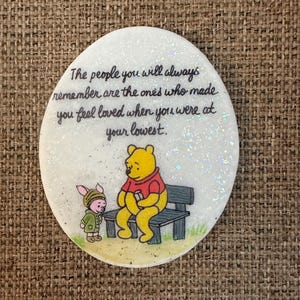May include: A round, white decorative item with a glittery surface. It features a Winnie the Pooh illustration with Piglet and a bench. The text reads: "The people you will always remember are the ones who made you feel loved when you were at your lowest."