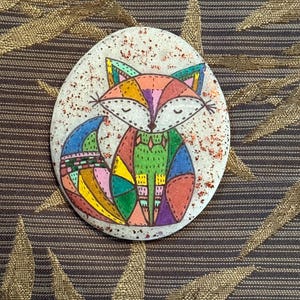 May include: A hand-painted, oval-shaped stone featuring a colorful fox design. The fox is depicted with geometric shapes in shades of orange, pink, green, blue, and purple. The stone has a speckled, off-white background with red glitter accents.