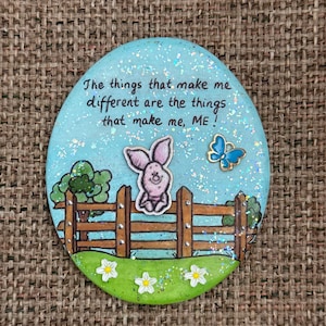 May include: A circular, light blue decorative item with glitter and a cartoon piglet. It features a wooden fence, flowers, and a butterfly. The text reads: "The things that make me different are the things that make me, ME!"