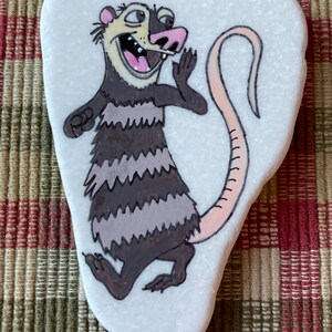 May include: Hand-painted rock art of a cartoon opossum with a pink nose and tail, brown and gray fur, and a wide open mouth. The opossum is painted on a white, oval-shaped rock.