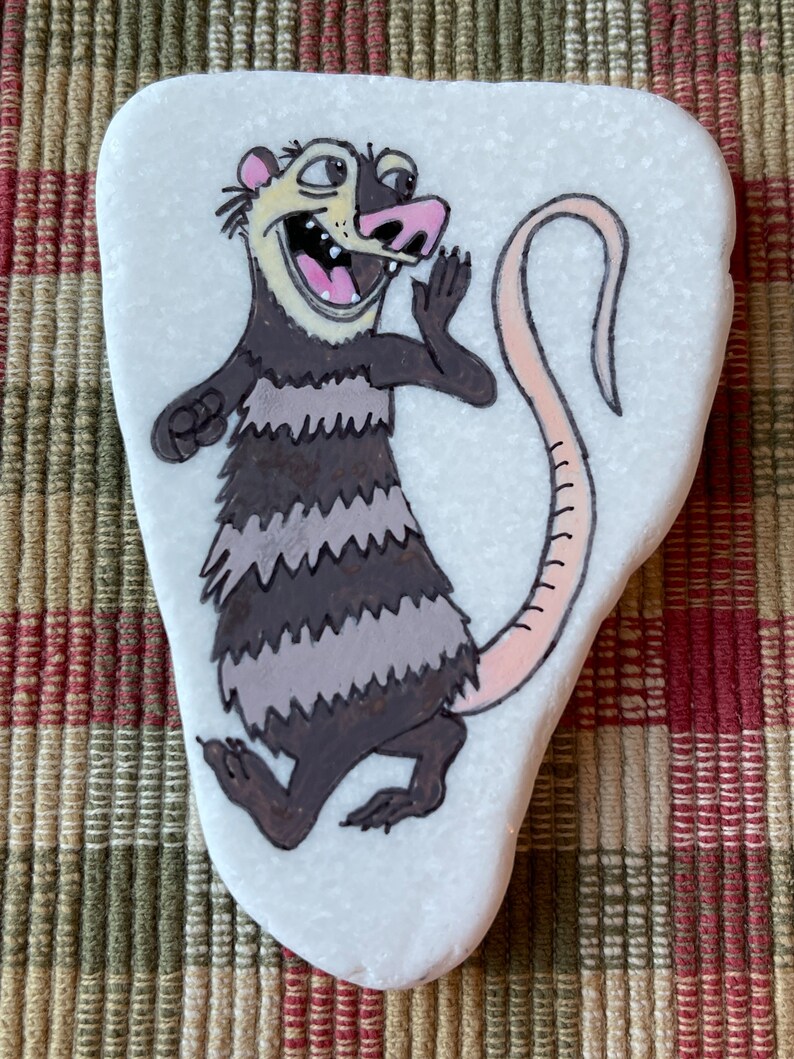 May include: Hand-painted rock art of a cartoon opossum with a pink nose and tail, brown and gray fur, and a wide open mouth. The opossum is painted on a white, oval-shaped rock.
