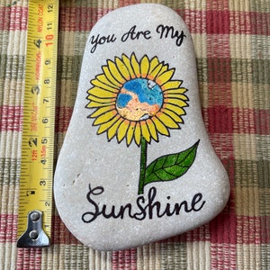 Puede incluir: Una piedra pintada con un dise&ntilde;o de girasol. El girasol tiene un centro amarillo con un globo azul y naranja en su interior. La piedra est&aacute; pintada con las palabras "You Are My Sunshine".