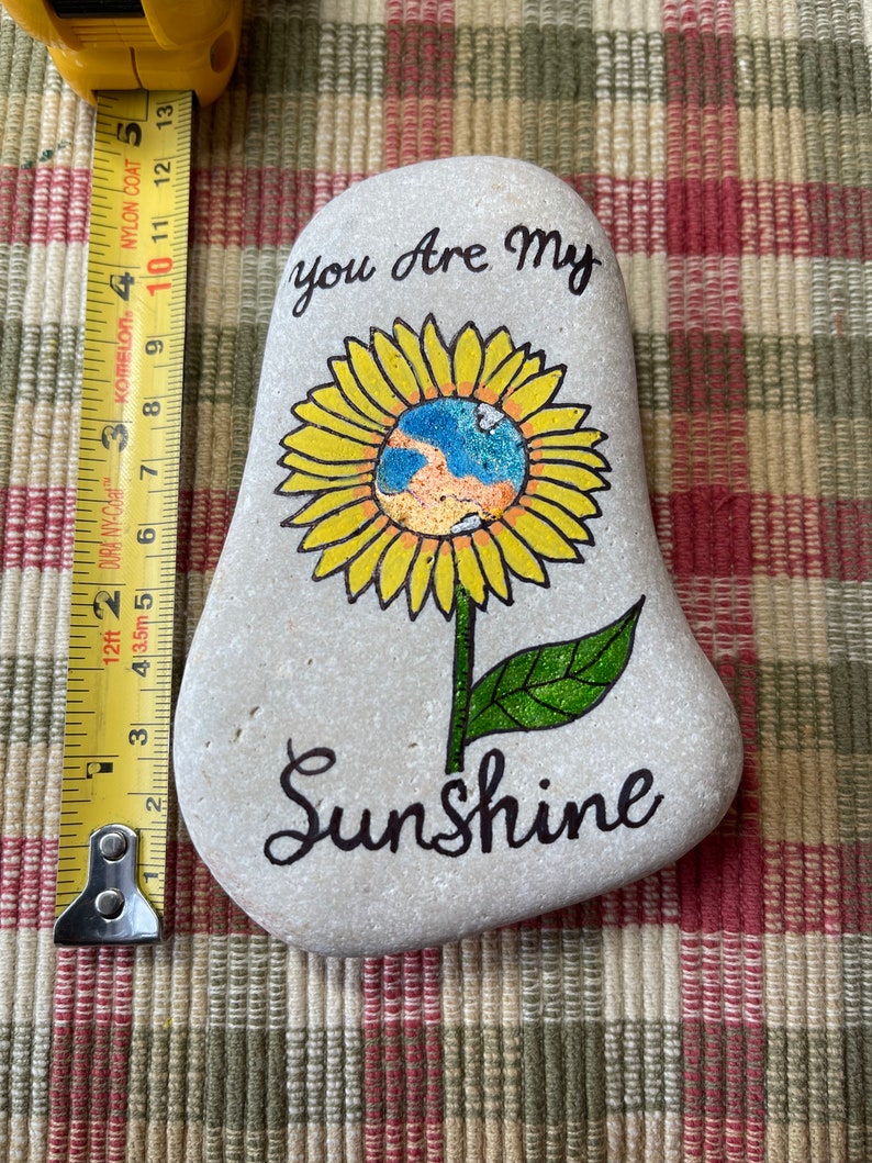 Puede incluir: Una piedra pintada con un dise&ntilde;o de girasol. El girasol tiene un centro amarillo con un globo azul y naranja en su interior. La piedra est&aacute; pintada con las palabras "You Are My Sunshine".