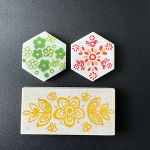 May include: Three decorative ceramic tiles with floral designs. Two hexagonal tiles feature green and red floral patterns. A rectangular tile displays a yellow floral design on a cream background. The tiles are arranged on a black surface.