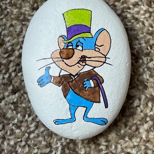 Puede incluir: Piedra pintada a mano con un rat&oacute;n de dibujos animados que lleva un sombrero de copa verde y morado y una chaqueta marr&oacute;n. El rat&oacute;n es azul con la nariz y los bigotes marrones.