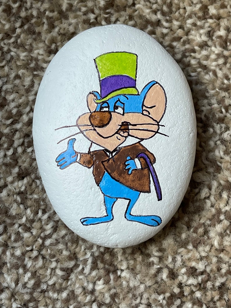 Puede incluir: Piedra pintada a mano con un rat&oacute;n de dibujos animados que lleva un sombrero de copa verde y morado y una chaqueta marr&oacute;n. El rat&oacute;n es azul con la nariz y los bigotes marrones.